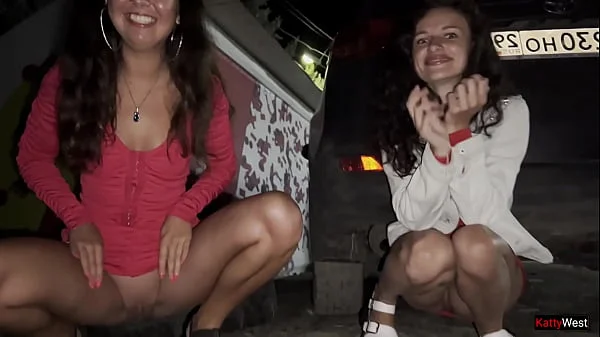 Two girlfriends pee together near a car in a public parking lot